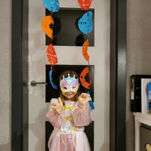 Ein Mädchen steht vor einer Tür, in einem Prinzessinnenkleid mit einer Maske auf. 